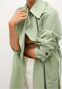 Jasnozielony płaszcz trench z szerokim kołnierzem i podwiniętymi rękawami, z guzikowymi wykończeniami i miękką, gładką fakturą. Noszony na beżowej bluzce.