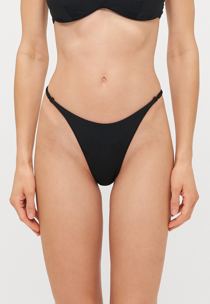 Fekete bikini alsó minimalista dizájnnal, vékony pántokkal és sima textúrával. Magasan kivágva a csípőnél a karcsú sziluettért.