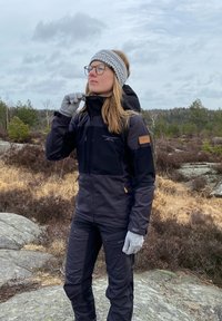 Zwarte waterdichte jas en broek met grijze handschoenen en hoofdband; kenmerken zijn zwarte en tan accenten, ritszakken en een snug pasvorm.