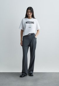 Tricou alb din bumbac cu imprimeu negru "Moschino Milano", asortat cu pantaloni gri eleganți, având o curea asortată. Încălțăminte neagră.