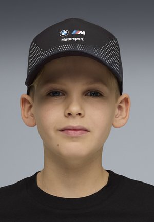 Chłopiec w czapce BMW Motorsport w kolorze czarnym z białymi kropkami oraz czarnej koszulce, na tle gładkiego, szarego tła.