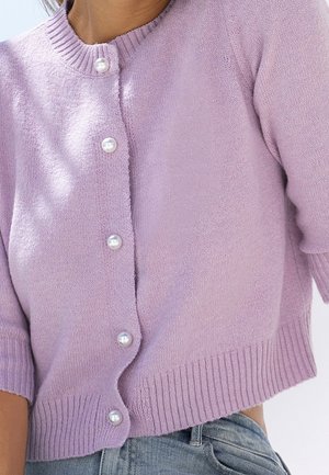 Kobieta ubrana w liliową dzianinową kardigan z perłowymi guzikami i prążkowanymi mankietami, w połączeniu z jasnoniebieskimi dżinsami.