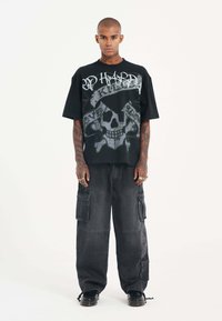 Camiseta negra de gran tamaño con un diseño gráfico de calavera, combinada con pantalones cargo gris oscuro con bolsillos. Hecha de tela de algodón.