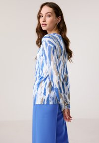 Leichtes Langarmoberteil mit einem blauen und beigen abstrakten Muster, mit einem runden Ausschnitt und gerippten Bündchen, kombiniert mit blauen Hosen.