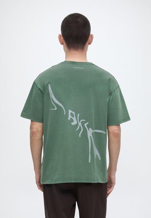 Άνδρας με κοντά σκουρόχρωμα μαλλιά γυρισμένος ανάποδα, φοράει ένα πράσινο φαρδύ t-shirt με μεγάλο αφηρημένο ανοιχτό γκρι σχέδιο στην πλάτη.