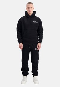 Zwarte hoodie met voorzak en logo op de borst, gecombineerd met bijpassende zwarte joggers. Beide gemaakt van zacht materiaal. Witte sneakers erbij.