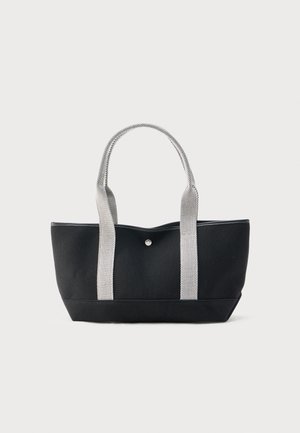 Svart totebag med brede hvite teksturerte håndtak og én sentral knappelukking, vist mot en enkel lys bakgrunn.