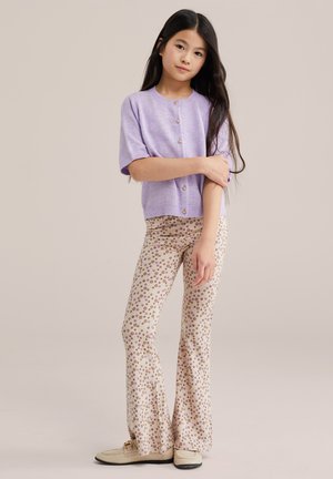 Jauna meitene ar gariem tumšiem matiem, valkājot gaiši violetu īsroku pogātu topu un bēša ziedu raksta izpletņbikses, stāv.