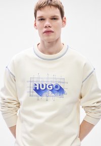 Krém színű pulóver kék "HUGO" grafikával és tervrajz dizájnnal. Bordázott mandzsettákkal és gallérral, puha tapintású, laza szabású.