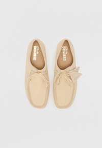 Scarpe mocassino beige in tessuto morbido, con punta rotonda, dettagli con lacci e un piccolo etichetta del marchio sul lato.