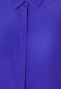 Živá fialová halenka s hladkou texturou, klasickým límcem a zapínáním na knoflíky. Má jednobarevný design bez viditelných vzorů.