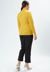 Suéter amarillo con cuello alto y puños acanalados, combinado con pantalones negros enrollados. El modelo lleva tacones de aguja plateados.