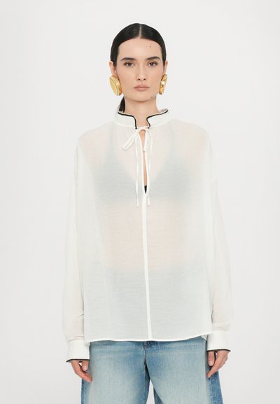 White, sheer long-sleeve blouse with a high collar, black trim, and a tie closure at the neck. Paired with light blue denim.