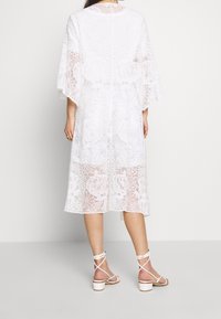 Abito midi in pizzo bianco con motivo floreale, texture semi-trasparente e maniche larghe. Abbinato a sandali bianchi con tacco a blocco e cinturini.