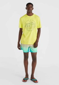 Camiseta amarilla neón con estampado gráfico, combinada con bañadores en degradado azul-verde con rayas horizontales. Sandalias negras.