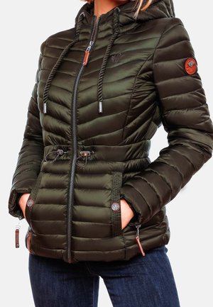 Femme portant une veste matelassée vert foncé avec capuche, fermeture éclair à l'avant, taille ajustable avec cordon, les mains dans les poches latérales, associée à un jean bleu.