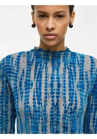 Robe à motifs bleus avec un col montant et des bords froncés. Le tissu semble texturé avec des nuances variées de bleu sur un fond clair.