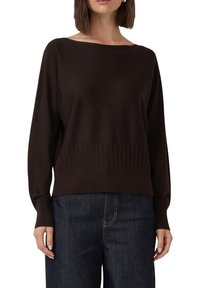 Femme portant un pull à manches longues marron foncé avec un ourlet et des poignets côtelés, associé à un jean en denim bleu foncé.