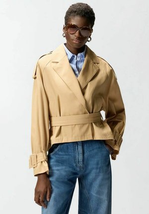 Donna con capelli corti indossa una giacca beige oversize con cintura, camicia a righe blu, jeans, occhiali da sole e orecchini a cerchio su sfondo semplice.