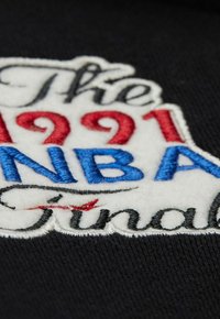 Tessuto nero con un design ricamato che presenta "Le Finali NBA 1991" in fili rossi, blu e bianchi, mostrando stili di testo variabili.