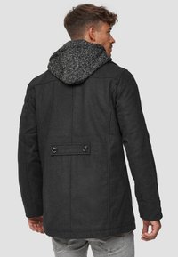 Svart ullkåpe med en teksturert grå hette. Har en bakventil og to knapper. Passformen er strukturert med en rett design.