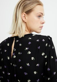Robe noire avec un imprimé floral en violet, blanc et vert. Fabriquée en tissu léger avec une ouverture en forme de clé dans le dos.