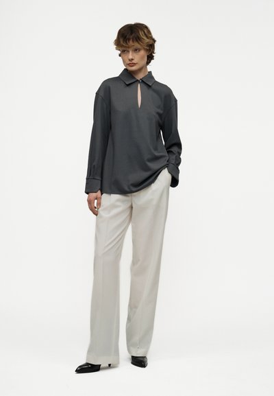 Άτομο που φοράει σκούρο γκρι πουκάμισο με μακριά μανίκια και γιακά με άνοιγμα στην κορυφή, και άσπρο παντελόνι με φαρδιά μπατζάκια, στέκεται με το ένα χέρι στην τσέπη.