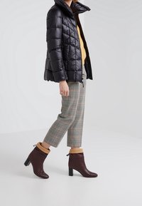 Personne portant une veste matelassée noire, un pantalon court à carreaux et des bottines marron avec des poignets couleur châtaigne, marchant sur un fond blanc uni.