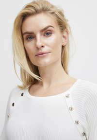 Bijela rebrasta puloverica s izrezanim dekolteom i dekorativnim gumbima uz jedno rame. Mekana tekstura, uski kroj.