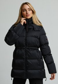 Giaccone puffoso nero con colletto alto, dettaglio in vita arricciato, chiusura frontale con zip e tasche laterali. Presenta un'etichetta con logo sulla manica.