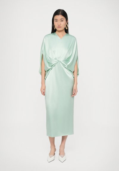 Γυναίκα που φοράει ένα γυαλιστερό μέντα πράσινο σατέν μεσάτο φόρεμα με ριγμένο εμπρός και μανίκια με σχισμή, συνδυασμένο με λευκά μυτερά τακούνια και μεγάλα χρυσά σκουλαρίκια κρίκους.