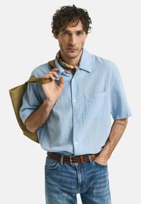 Camicia azzurra a maniche corte con tessuto testurizzato e taschino, abbinata a jeans blu e una cintura intrecciata marrone.