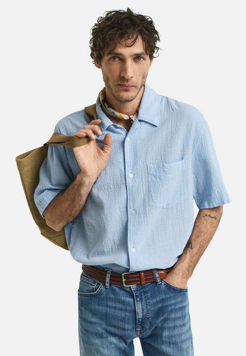 Camicia azzurra a maniche corte con tessuto testurizzato e taschino, abbinata a jeans blu e una cintura intrecciata marrone.
