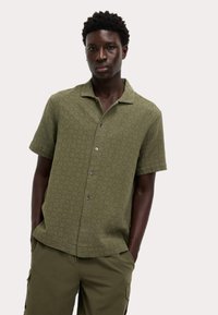 Camisa de manga corta de color verde oliva con un patrón geométrico texturizado, cierre de botones y cuello clásico. Combinada con unos pantalones cortos cargo a juego.