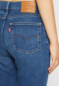 Jeans de mezclilla azul que presentan un diseño distintivo en el bolsillo trasero, una etiqueta de marca de cuero y acentos en rojo con costuras naranjas.