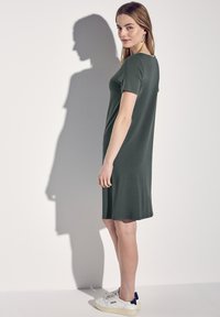 Ciemnozielona sukienka z krótkim rękawem o luźnym kroju i długości do kolan, zestawiona z białymi trampkami, podkreślona subtelnym cieniem.