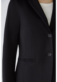 Personne portant un blazer noir ajusté à deux boutons sur un haut blanc, montrée de l'épaule à la taille sur un fond uni.