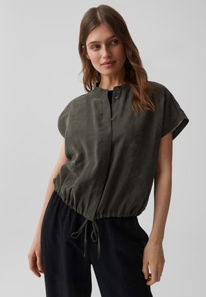 Femme portant une chemise boutonnée manches courtes olive foncé avec taille à cordon et pantalon noir ample, debout devant un fond gris uni.