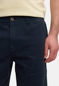 Pantalons en coton bleu marine avec fermeture à bouton et poches latérales, présentant une texture subtile et une étiquette de marque près de la taille.
