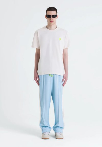 T-shirt di cotone chiaro con piccolo logo verde, abbinato a pantaloni da jogging di satin blu chiaro con strisce laterali e cordoncino.