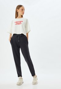 Witte cropped t-shirt met rode tekst "BETER DAN VESTERDAG," gecombineerd met zwarte joggers met zijzakken en grijze sneakers.