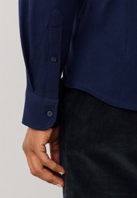 Marineblauw overhemd met lange mouwen, gemaakt van een gestructureerde stof en met twee knopen op de manchetten en een licht gebogen zoom. Hand zichtbaar die de rand van het shirt vasthoudt.