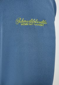 Tessuto blu testurizzato con testo ricamato verde che recita "Schmuddelwedda WENN DAT FLADDELT."