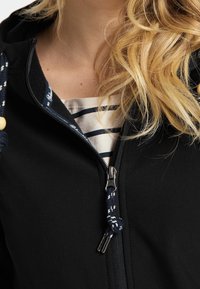 Svart huva jacka med dragkedja fram. Har ett mönstrat snöre i blått och vitt och visar en randig skjorta under.