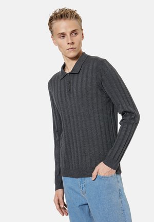 Młody mężczyzna w ciemnoszarym prążkowanym swetrze polo z długim rękawem i jasnoniebieskich dżinsach, patrzący w lewo z jedną ręką w kieszeni.