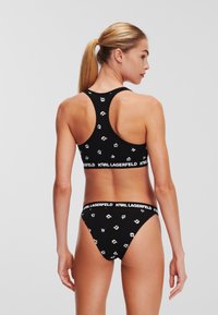 Fekete bikini szett, amely racerback bralette-ből és alacsony derékú alsóból áll, ismétlődő logó mintával díszítve és márkás rugalmas derékpánttal.