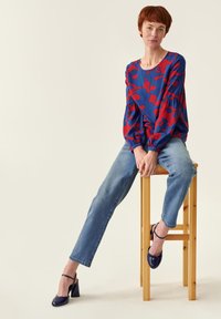 Blusa de estampado rojo y azul con mangas abullonadas, combinada con jeans de color azul claro y zapatos de tacón alto en tono marino, sentada en un taburete de madera.