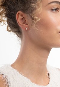 Boucle d'oreille en argent avec une petite pierre ronde claire, sertie dans un simple chaton. Des cheveux bouclés encadrent l'oreille, mettant en valeur les détails de la boucle d'oreille.