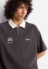Polo gris oscuro con un cuello en contraste de color crema. Presenta un logo bordado en el pecho y en la manga izquierda. Tejido suave y texturizado.