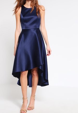 Femme portant une robe bleu marine en satin sans manches à ourlet asymétrique, avec des talons nus à bride à la cheville, debout devant un fond blanc uni.